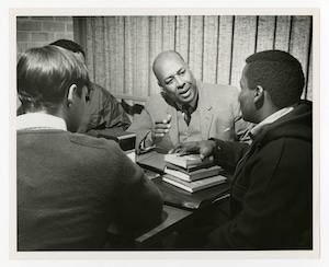 Mr. Frank Hercules speaking with students seated around him and one with his hand on a stack of books